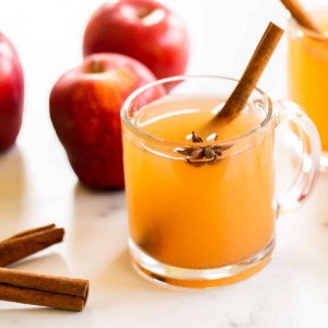 A marble surface with clear mugs full of homemade apple cider, whole apples and cinnamon sticks to the side.