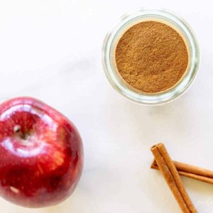 A marble surface with a small jar of apple pie spice, cinnamon sticks and an apple to the side.