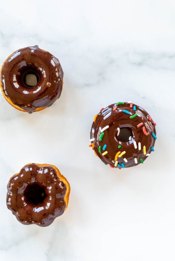 Baked donuts topped with chocolate frosting and sprinkles, on a marble countertop.