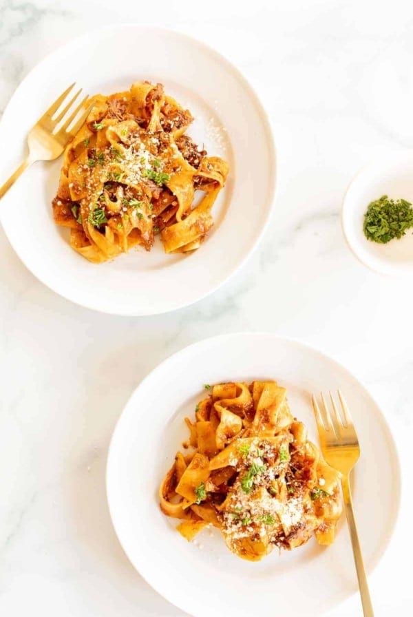 Two white plates on a marble surface, filled with a beef ragu recipe and a small bowl of parsley to the side.