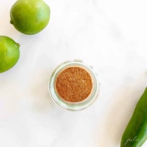 Marble surface with a clear jar full of fajita seasoning, limes and jalapeno to the side.