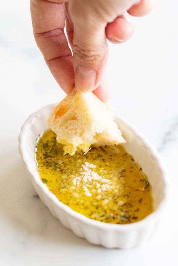 A hand dipping a piece of bread into a white ramekin full of olive oil bread dip.