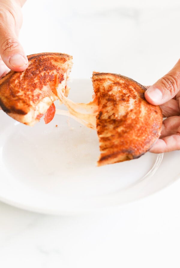 Hands pulling apart a campfire pizza sandwich with melted cheese on a white plate.