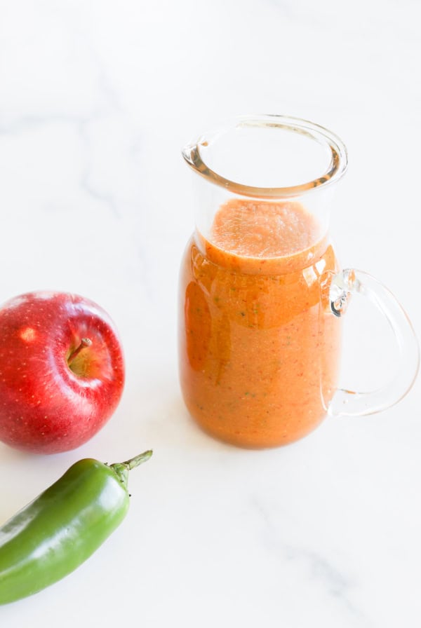 A glass pitcher filled with orange smoothie, accompanied by a red apple and a green chili pepper on a white marble surface, next to a bowl of chipotle salad dressing.