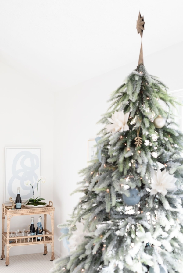 A frosted Christmas tree decorated with white flowers and ornaments evokes a Coastal Christmas feel as it stands beside a small wooden cart holding wine bottles and glasses in a bright room.