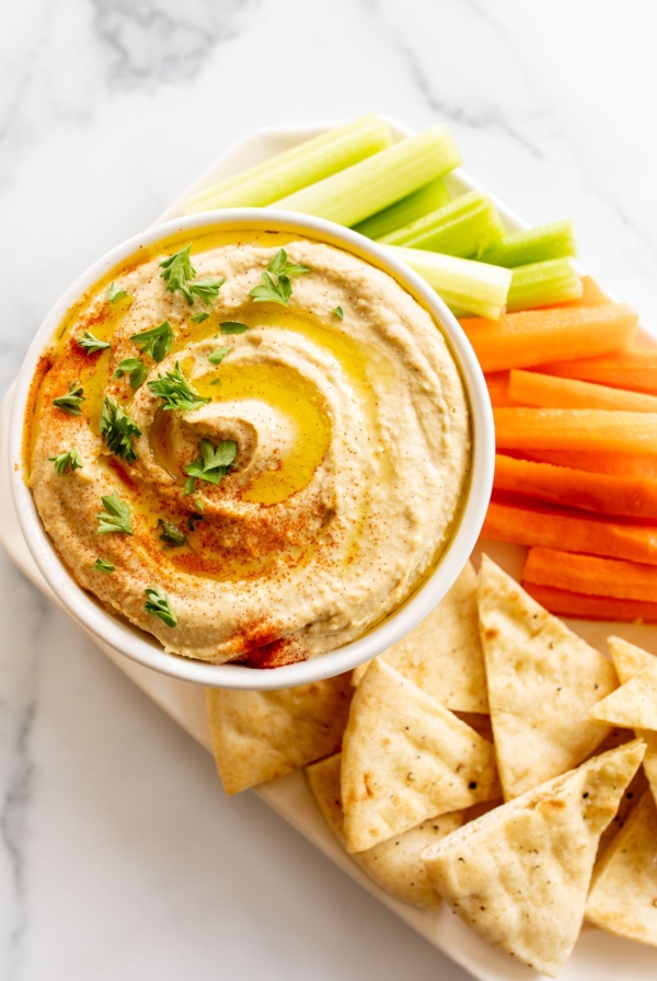 A white bowl of hummus surrounded by a platter of vegetables for dipping.