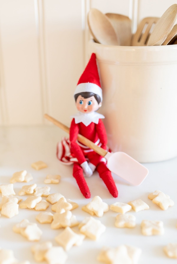 An elf on the shelf sitting on a kitchen counter, with tiny shortbread elf on the shelf cookies surrounding her.