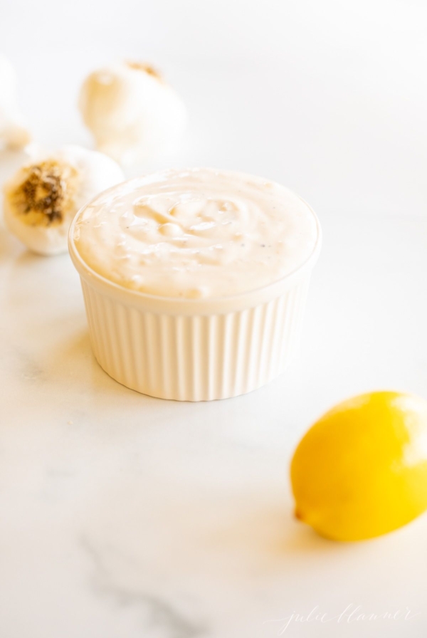 A white ramekin full of garlic aioli with a garlic bulb and a lemon in the foreground.