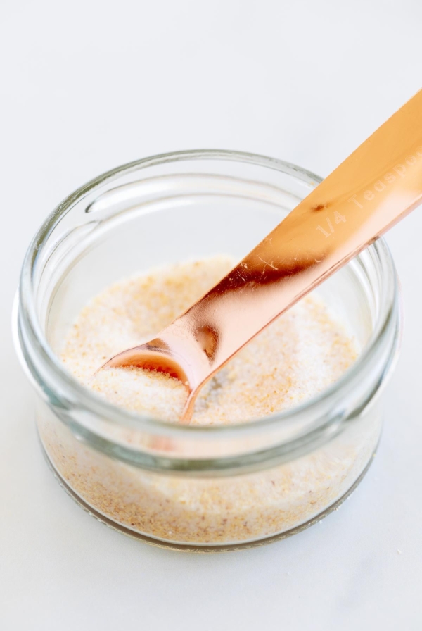 A small glass jar filled with homemade garlic salt, placed on a marble countertop. A copper measuring spoon is in the jar.