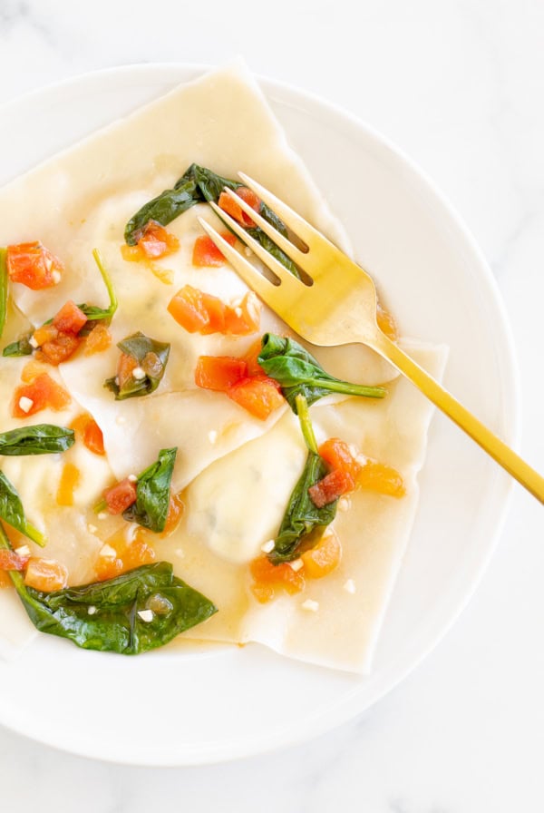 A plate of goat cheese ravioli topped with chopped tomatoes and spinach leaves, served on a white marble background.