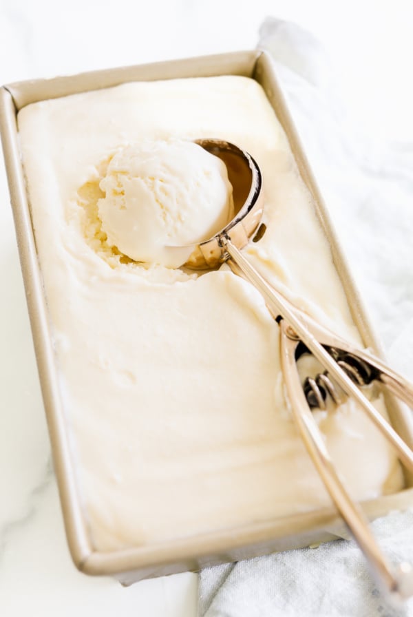 A metal ice cream scoop rests in a rectangular container of Gooey Butter Cake ice cream, with one scoop of the rich treat inside the scooper. A white cloth is partially visible beside the container.