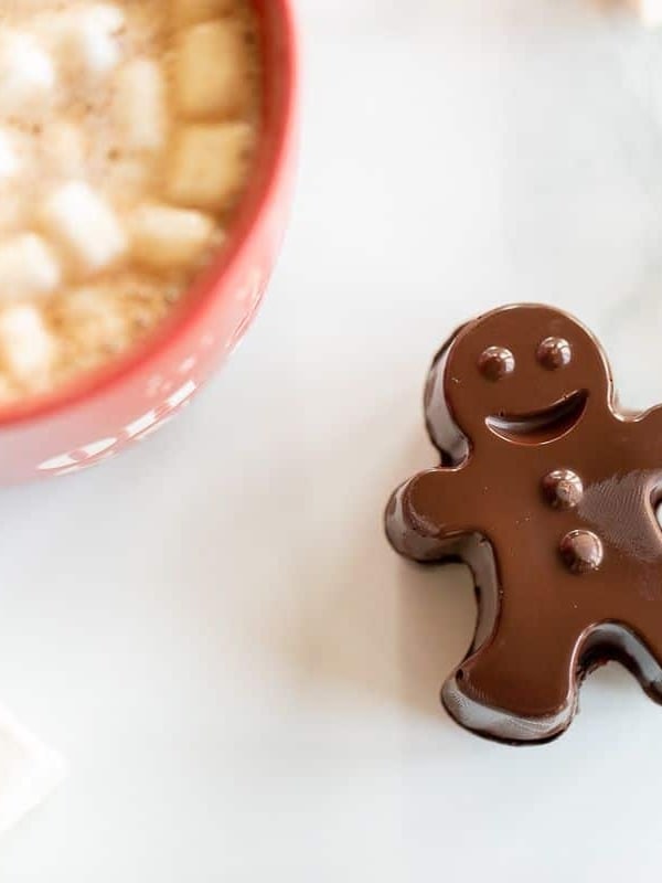 A red mug full of dark hot chocolate with marshmallows, with a dark chocolate gingerbread man to the side.