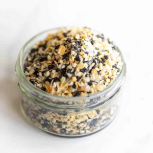 A clear glass jar on a marble surface, filled with everything bagel seasoning.