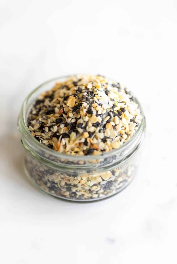 A clear glass jar on a marble surface, filled with everything bagel seasoning.