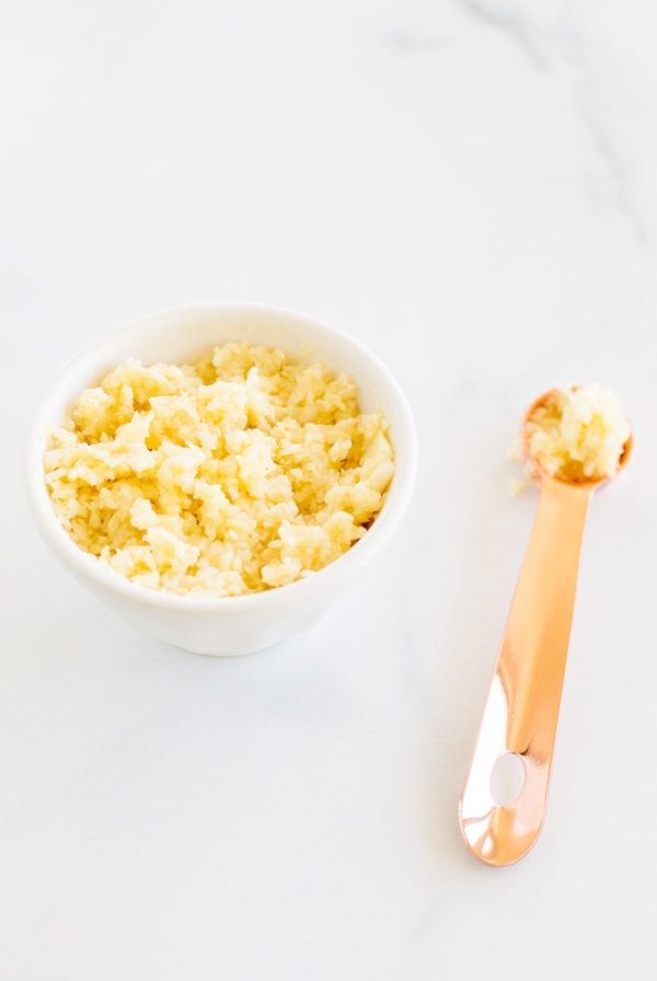 A small white bowl of minced garlic, with a copper teaspoon full of minced garlic next to it.