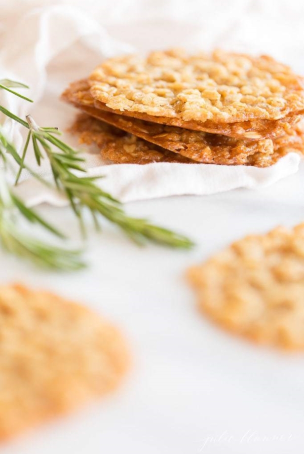 A stack of oatmeal lace cookies on a white surface, with a touch of greenery in the corner