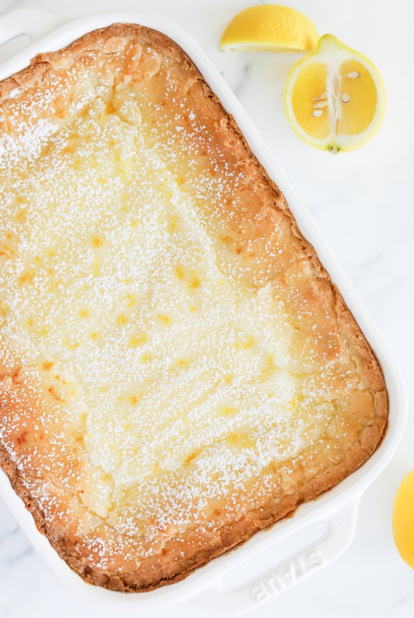 A Lemon Gooey Butter Cake sits elegantly in a rectangular white dish, dusted with powdered sugar to enhance its sweetness, with two fresh lemon halves on the side.