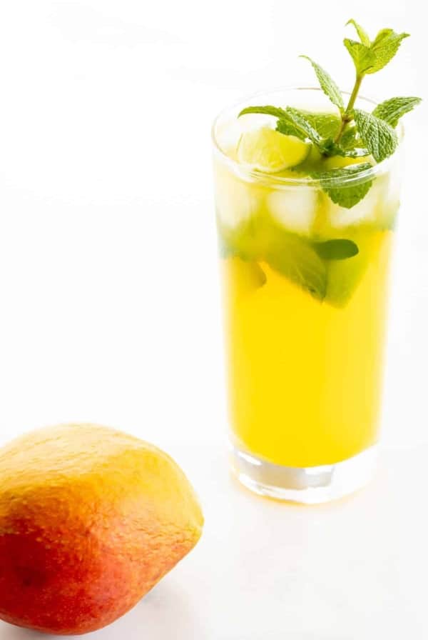 A mango mojito recipe in a clear glass, garnished with a sprig of mint, mango in foreground.