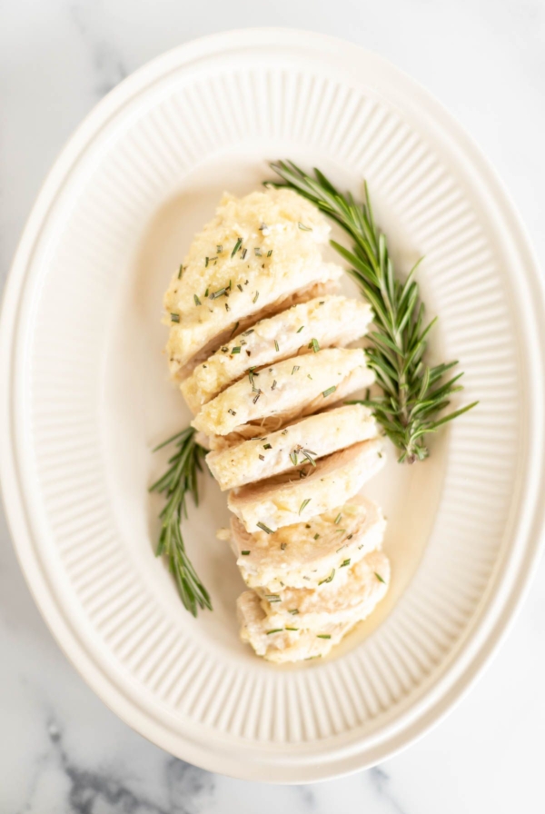 Sliced mayo chicken, garnished with fresh rosemary on an oval platter.