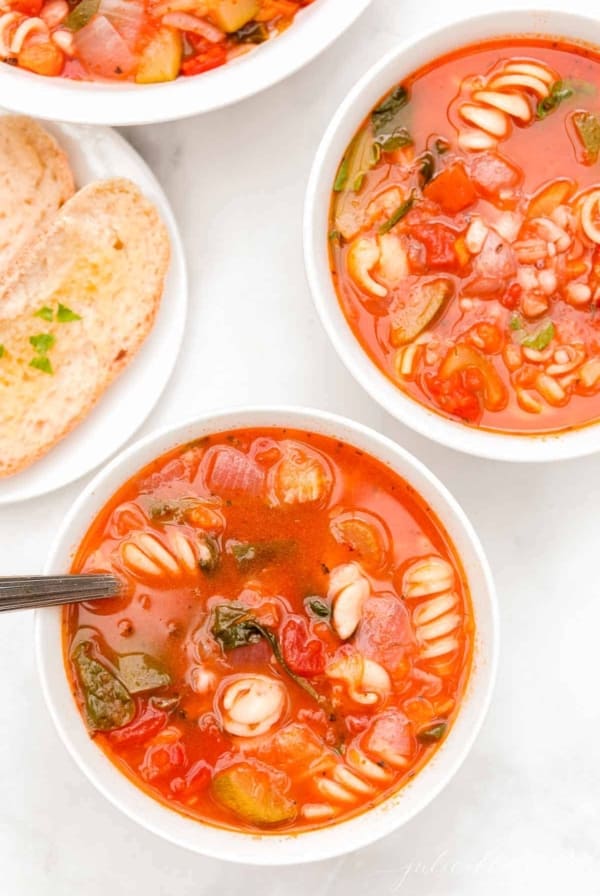 A white bowl of minestrone soup, another bowl in the background with herbs and a slice of bread on the surface.