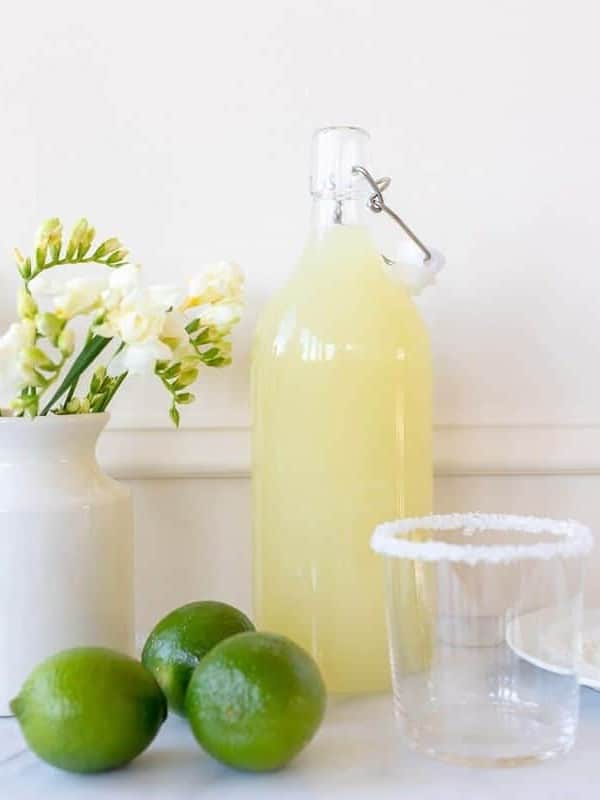 White kitchen background, glass carafe full of homemade margarita mix, limes to the side.