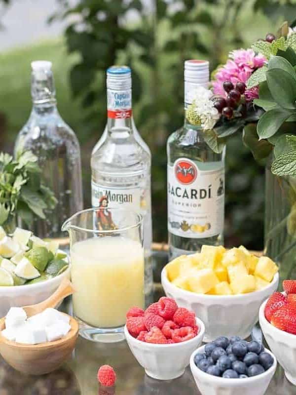 A glass and brass bar cart set up with a mojito bar outdoors, various fruits and limes in bowls beside alcohol bottles.