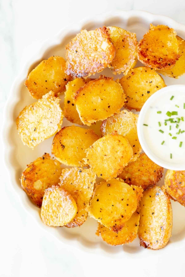 A white ruffled plate full of parmesan potatoes, with a small bowl of sour cream dip.