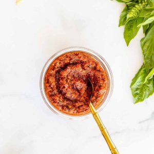 A small white bowl of pesto rosso, placed next to basil leaves, placed on a marble countertop.