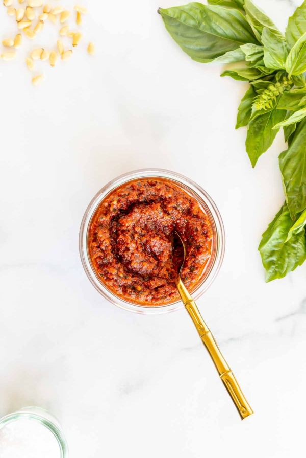 A small white bowl of pesto rosso, placed next to basil leaves, placed on a marble countertop.