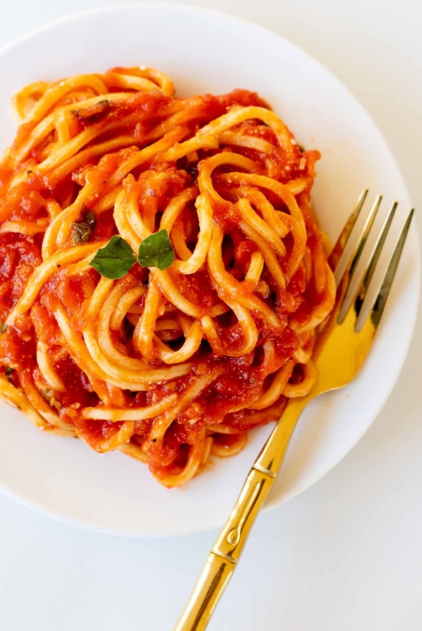 Spaghetti on a white plate, tossed in San Marzano tomato sauce, with a gold fork to the side.