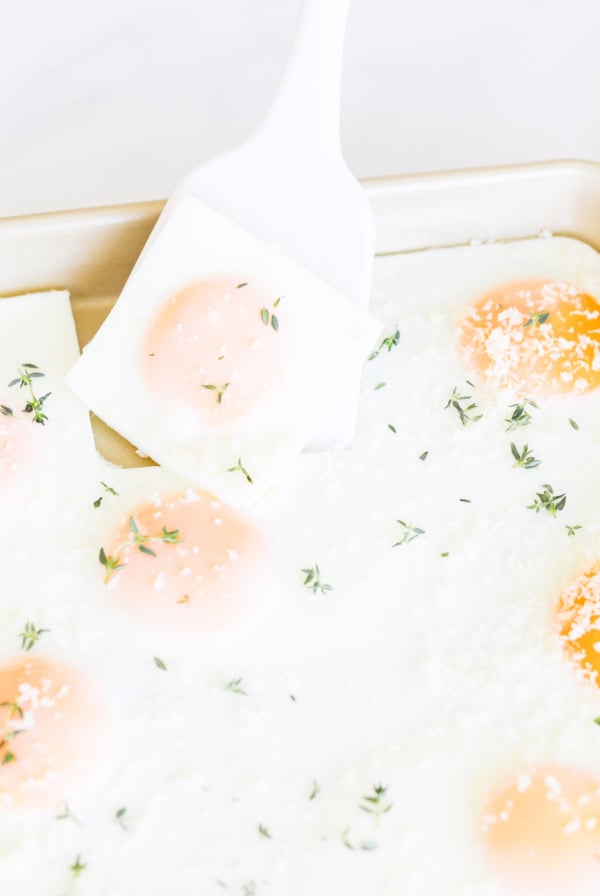 A spatula lifts a square portion of sheet pan eggs with a few sprigs of thyme and grated cheese from a tray.