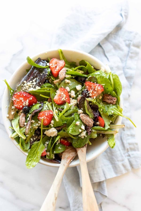 A fresh spring salad with berries, crumbled cheese and nuts in a bowl with wooden serving utensils.