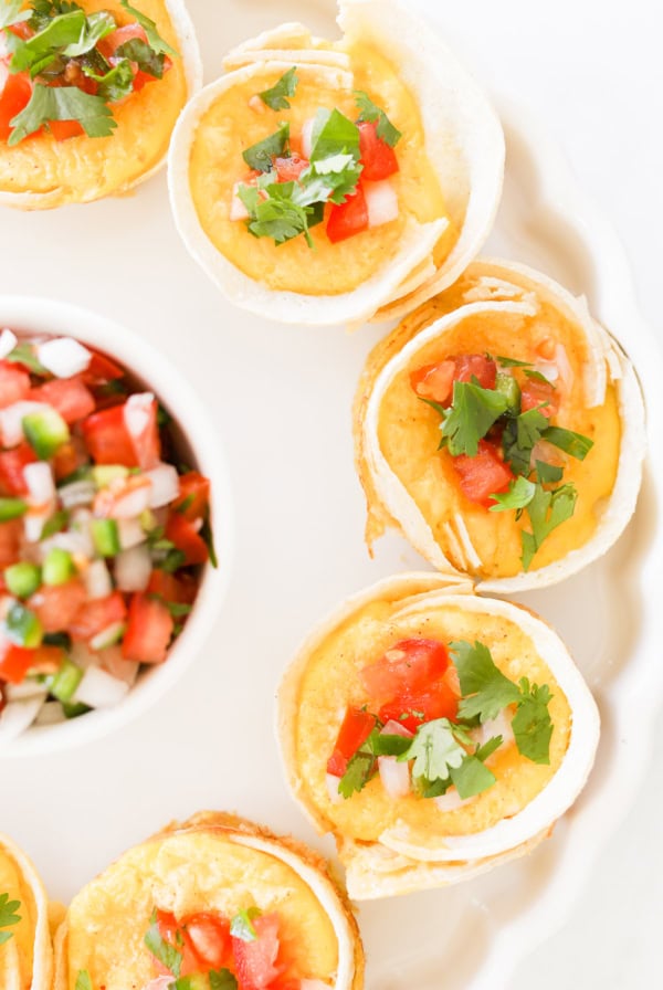 A plate of mini Mexican Breakfast Cups filled with melted cheese, diced tomatoes, onions, and cilantro, arranged in a circle with a small bowl of salsa in the center.