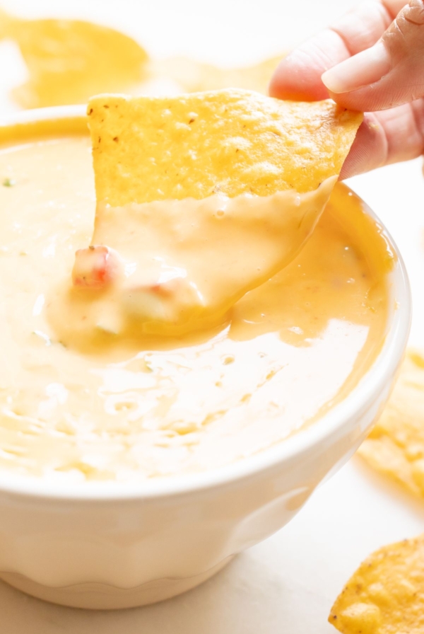 A hand dipping a tortilla chip into a white bowl of Velveeta cheese dip.