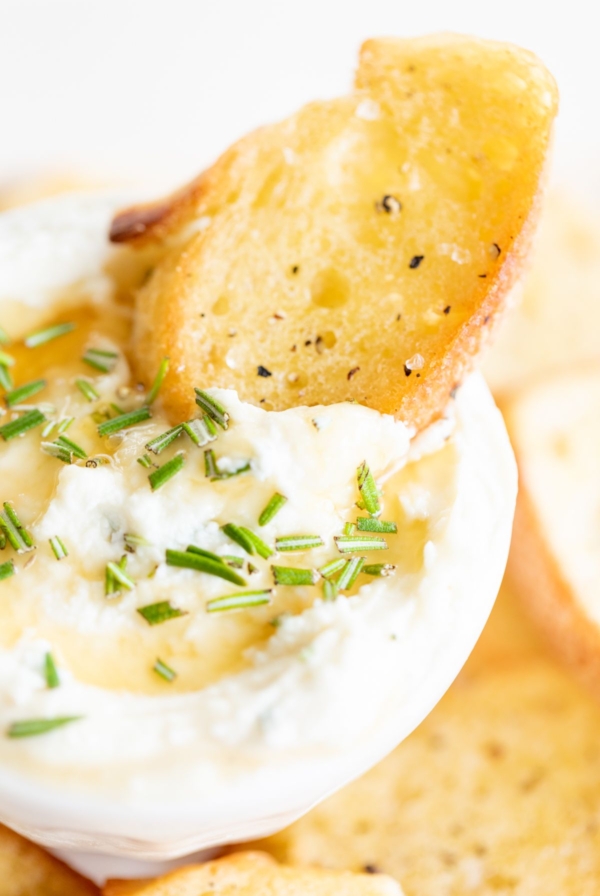 Whipped goat cheese with honey in a white bowl, on a platter of crostini for dipping.